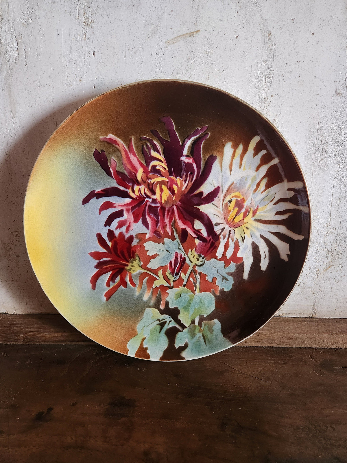 A round ceramic plate featuring a hand-painted floral design with chrysanthemums in red, white, and yellow, set against a dark brown background with a yellow gradient on the left, resting on a rustic wooden surface.