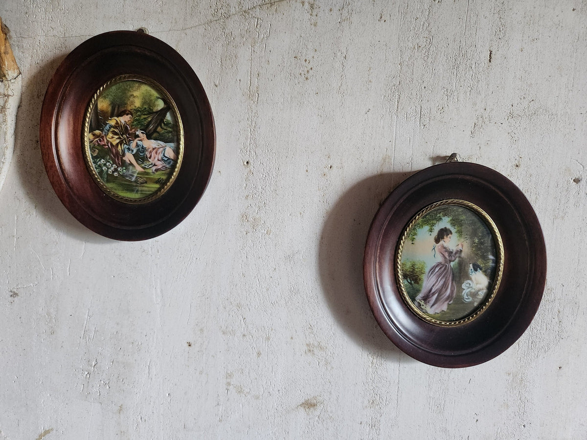 May include: Two oval framed pictures with brown wood frames and gold trim. The pictures are hanging on a white wall. The pictures contain paintings of people in a garden setting.