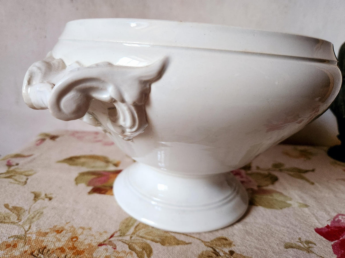 May include: A white ceramic serving bowl with a pedestal base. The bowl has a wide, rounded shape and features decorative handles with a sculpted design. The bowl is approximately 25 cm in diameter.