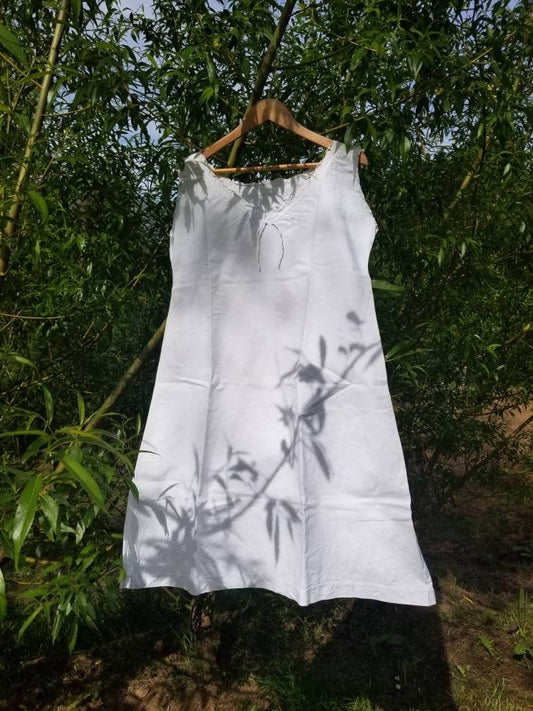 May include: A white cotton slip dress with a lace trim neckline hanging on a wooden hanger. The dress is hanging in front of a green leafy background.